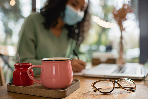 cup of coffee near sugar bowl and african american woman in medical mask on blurred background