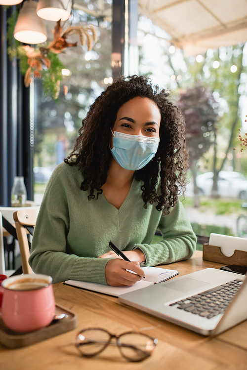 curly african american woman in medical mask writing in notebook near laptop in cafe