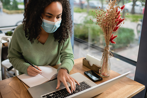 african american woman in medical mask using laptop near smartphone and notebook on table