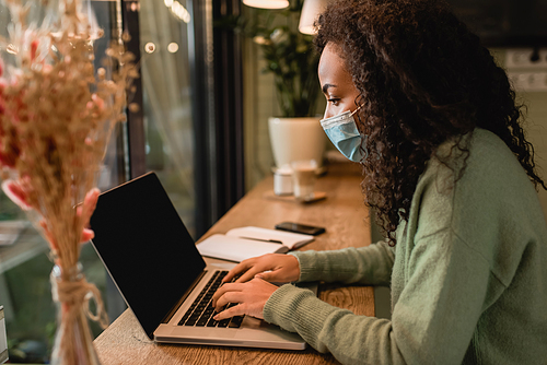 african american woman in medical mask using laptop with blank screen in cafe