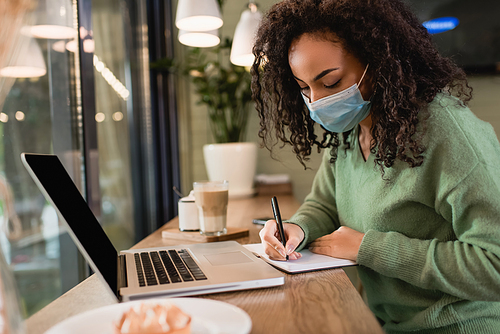 african american woman in medical mask writing in notebook near laptop with blank screen on table
