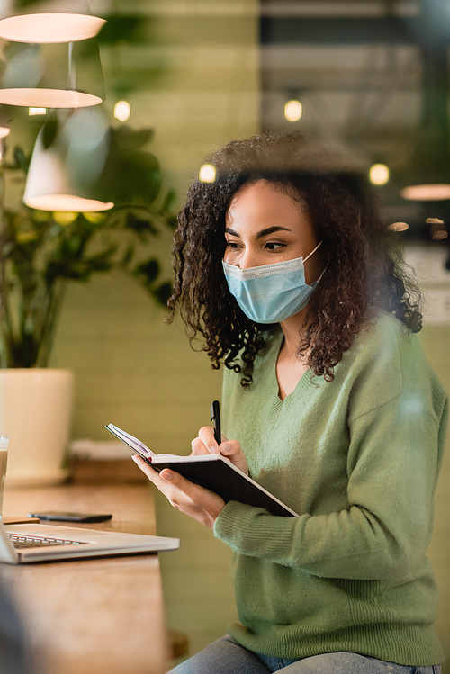 african american woman in medical mask holding notebook and pen while looking at laptop