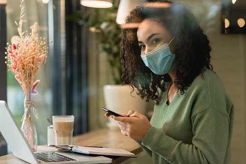 african american woman in medical mask holding smartphone near laptop and glass of latte in cafe