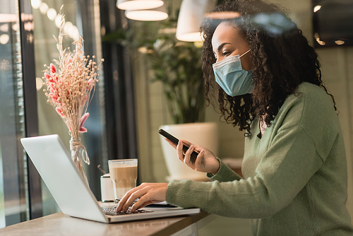 curly african american freelancer in medical mask holding smartphone near laptop and glass of latte in cafe