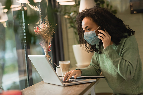 african american woman in medical mask talking on smartphone while using laptop near notebook on table