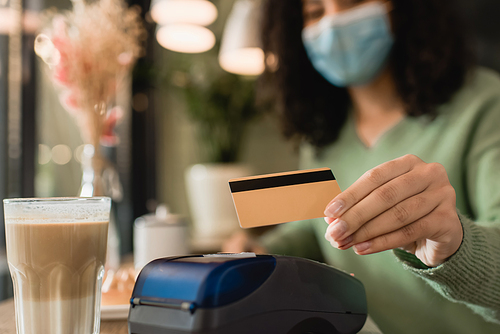 glass of latte near credit card in hand of african american woman in medical mask paying on blurred background
