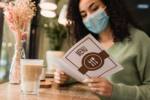 african american woman in medical mask reading menu near glass of latte in cafe