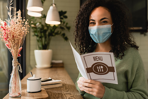 african american woman in medical mask holding brochure with menu lettering in cafe