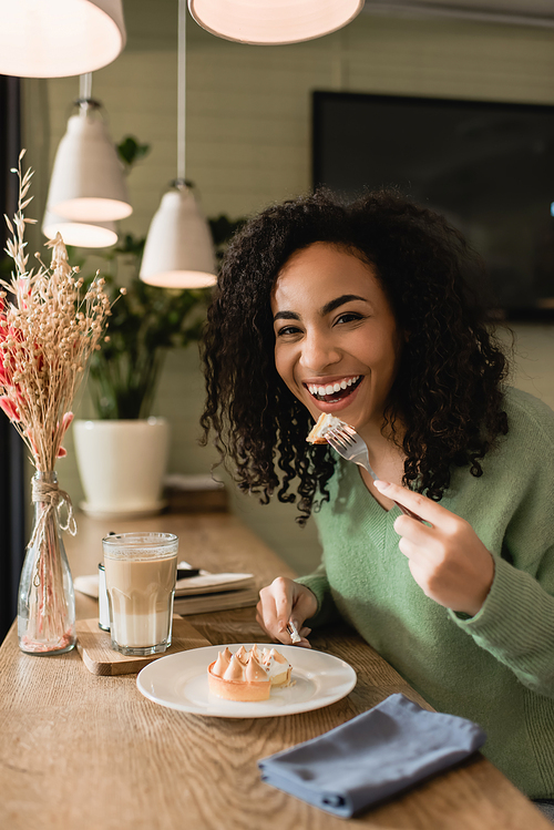 cheerful african american woman eating tasty tart near glass of latte in cafe