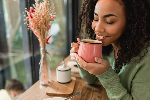 pleased african american woman holding pink cup with coffee in cafe