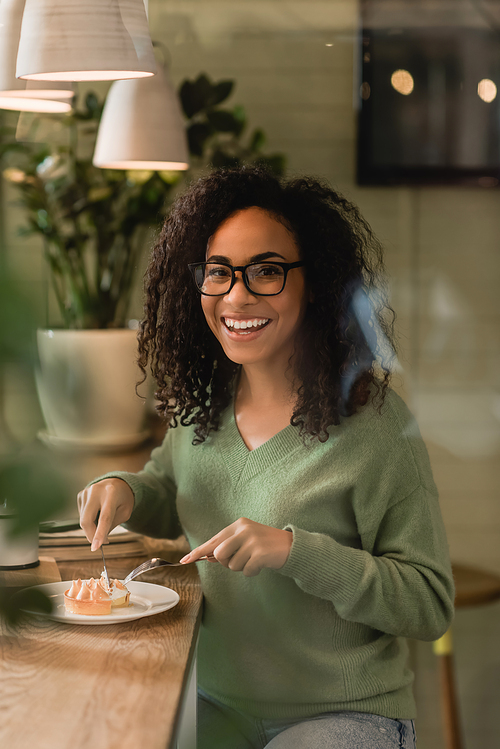 cheerful african american woman in eyeglasses holding cutlery near tart on plate