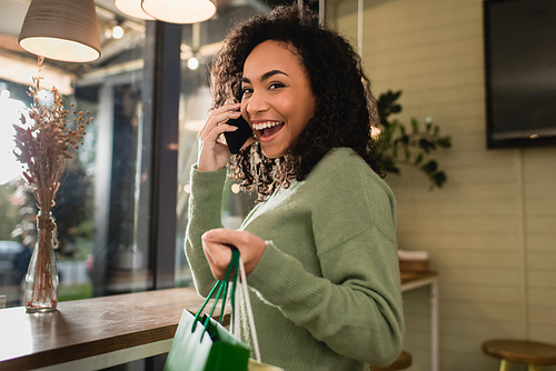 smiling african american woman talking on smartphone and holding shopping bags in cafe