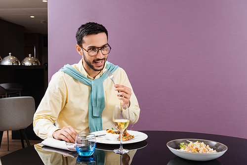 Smiling arabian man holding fork near meal and wine in hotel room