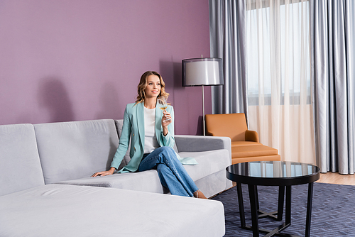 Smiling woman with glass of wine sitting on sofa in modern hotel room