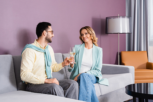 Smiling interracial couple with glasses of wine sitting on sofa in hotel room