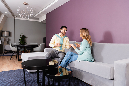 Cheerful muslim man with glass of wine holding hand of girlfriend in hotel room