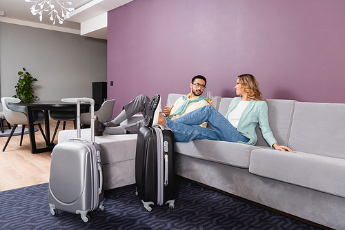 Suitcases near interracial couple holding glasses of wine in hotel room