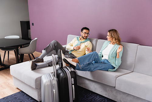 Interracial couple holding wine glasses near luggage in hotel