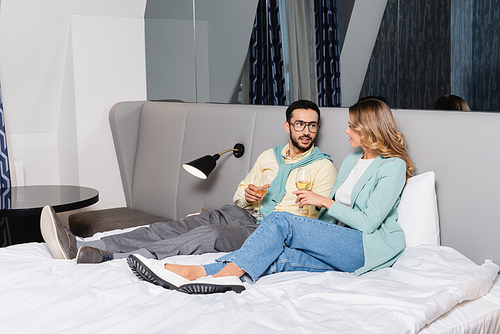 Muslim man with glass of wine looking at girlfriend on hotel bed