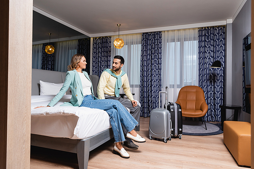 Smiling woman looking at muslim boyfriend on bed near suitcases in hotel