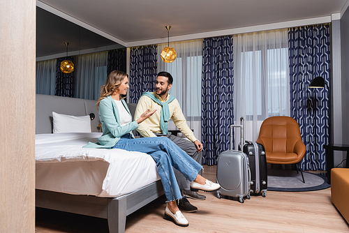 Smiling woman pointing with hand near suitcases and muslim boyfriend on bed in hotel