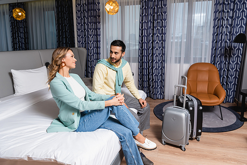 Smiling interracial couple sitting on bed near suitcases in hotel room