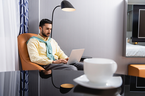 Muslim man using laptop on armchair near cup of coffee on blurred foreground in hotel room