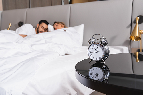 Alarm clock on table and sleeping interracial couple on blurred background in hotel