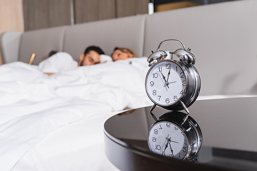 Alarm clock near sleeping couple on blurred background in hotel
