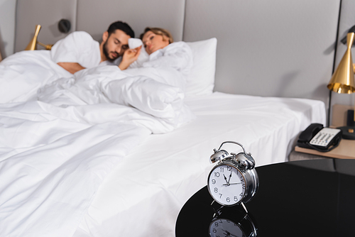 Alarm clock on table near couple sleeping on blurred background in hotel room