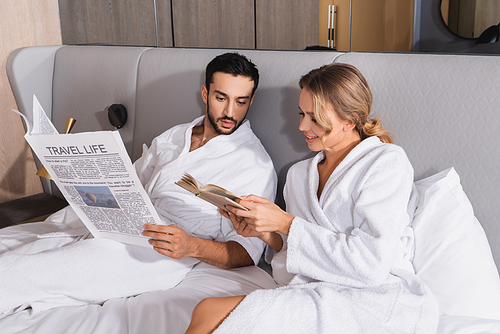 Smiling woman showing book to muslim boyfriend with newspaper on hotel bed