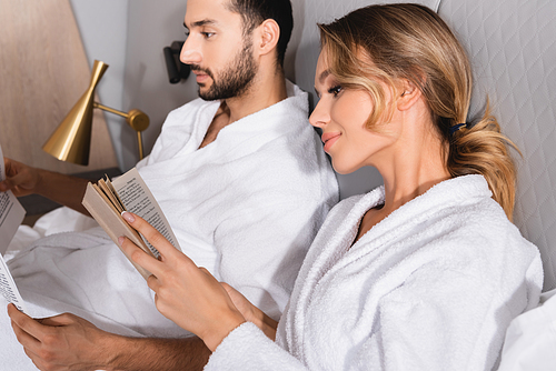 Young woman reading book near muslim boyfriend with news on blurred background in hotel