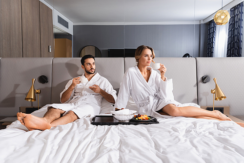 Interracial couple with cups sitting near breakfast on hotel bed