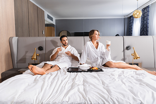 Young woman holding cup near breakfast and muslim boyfriend on hotel bed