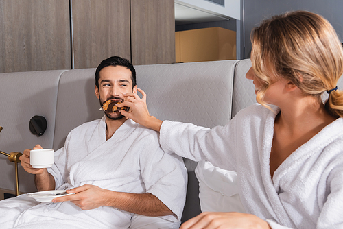 Young woman feeding croissant to arabian boyfriend with cup on bed in hotel