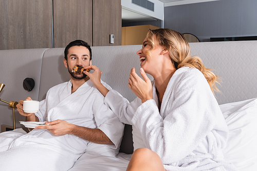 Cheerful woman feeding croissant to muslim boyfriend with coffee on hotel bed