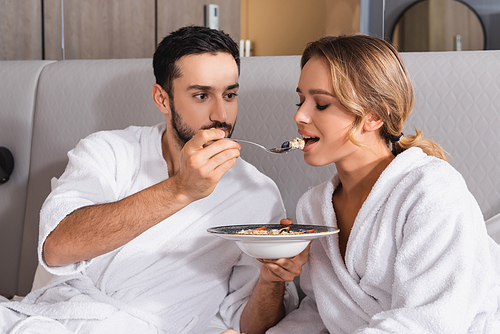 Arabian man in bathrobe feeding salad to girlfriend on hotel bed
