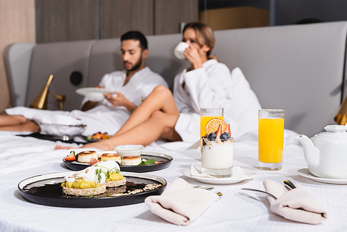 Delicious desserts and orange juice on bed near couple on blurred background in hotel room