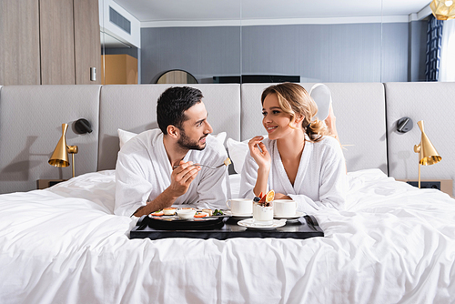 Smiling interracial couple looking at each other near breakfast on tray on hotel bed