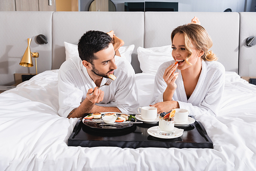 Young interracial couple in bathrobes eating breakfast near tray on hotel bed