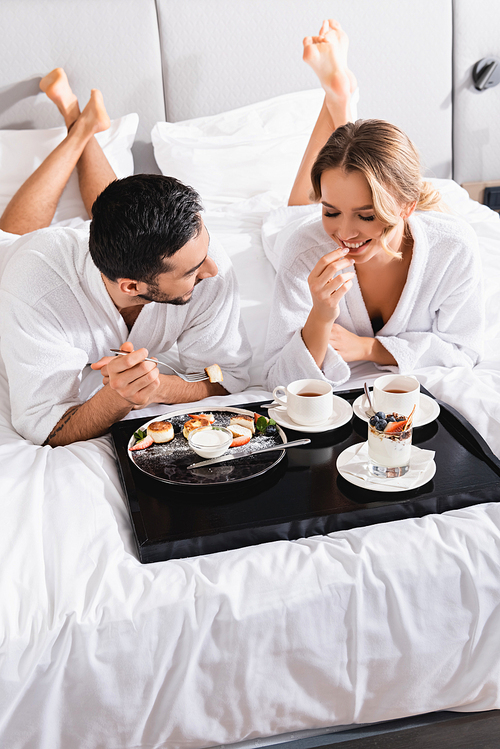 Smiling woman looking at desserts on tray near muslim boyfriend on hotel bed