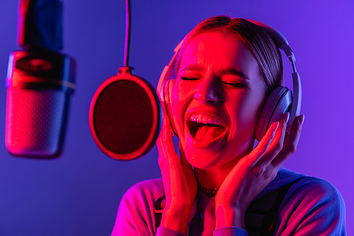 young singer in wireless headphones singing song in microphone on blurred foreground and purple