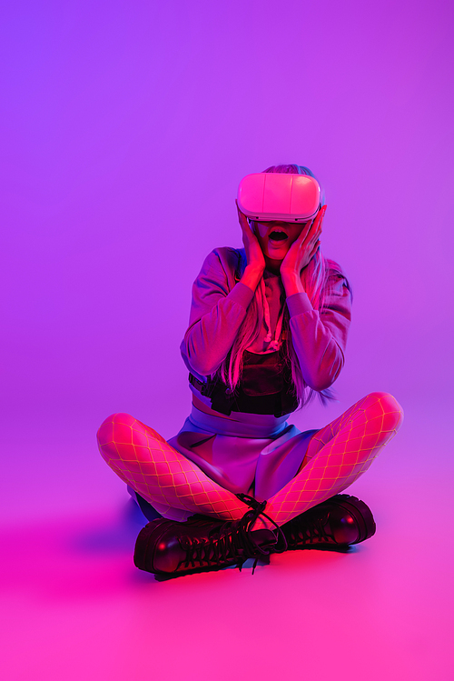 full length of afraid woman in virtual reality headset sitting on purple background