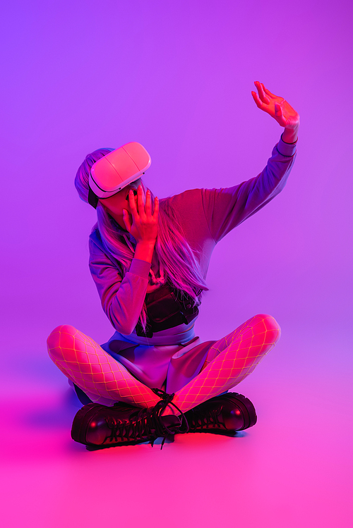 surprised woman in virtual reality headset sitting with crossed legs and covering mouth while gesturing on purple