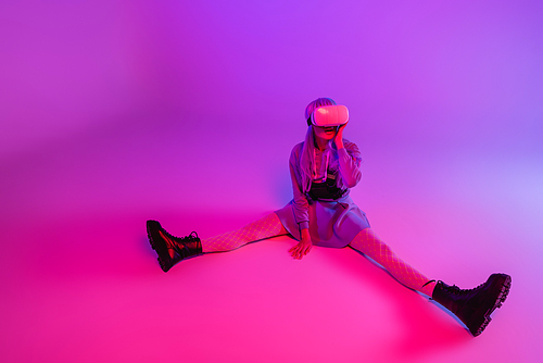 full length of flexible woman in virtual reality headset sitting on purple background with pink lighting