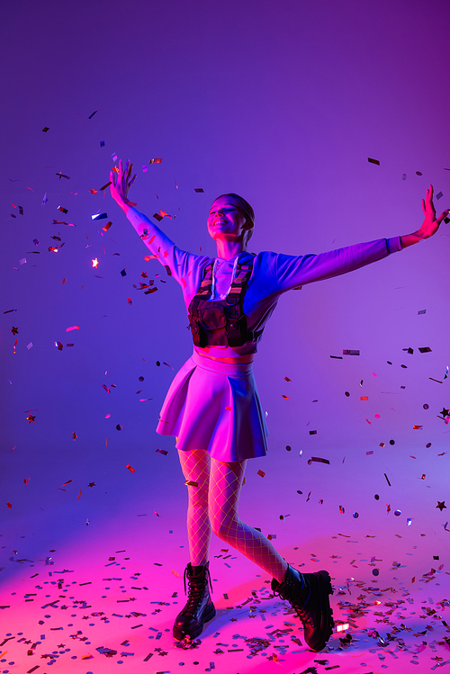 full length of happy woman in stylish outfit with outstretched hands near falling confetti on purple