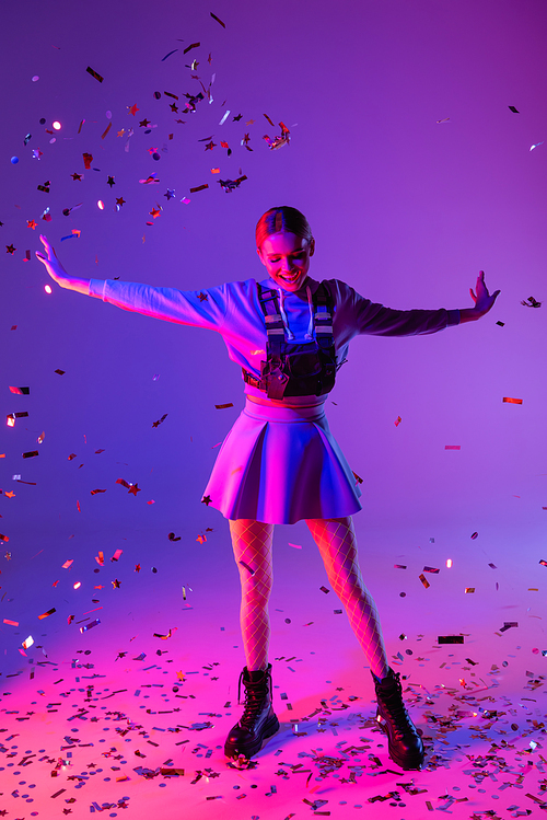 full length of cheerful woman in stylish outfit with outstretched hands near falling confetti on purple