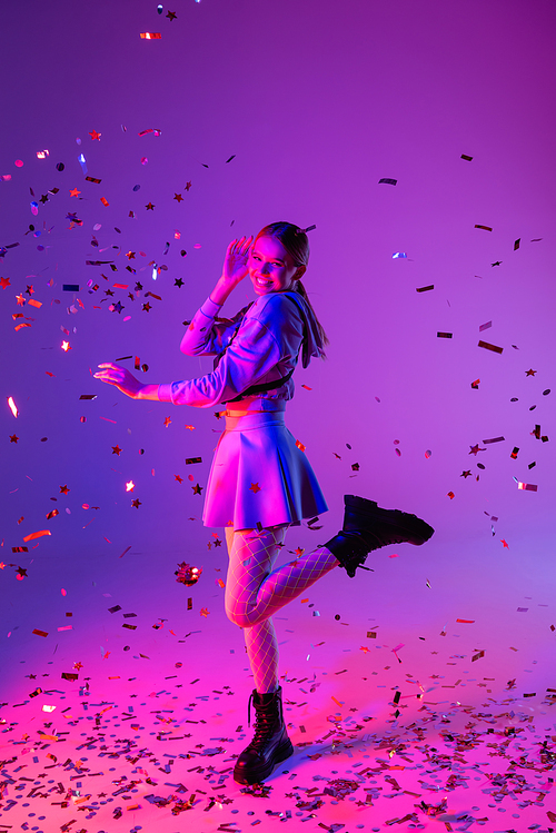 full length of happy woman in stylish outfit winking eye near falling confetti on purple