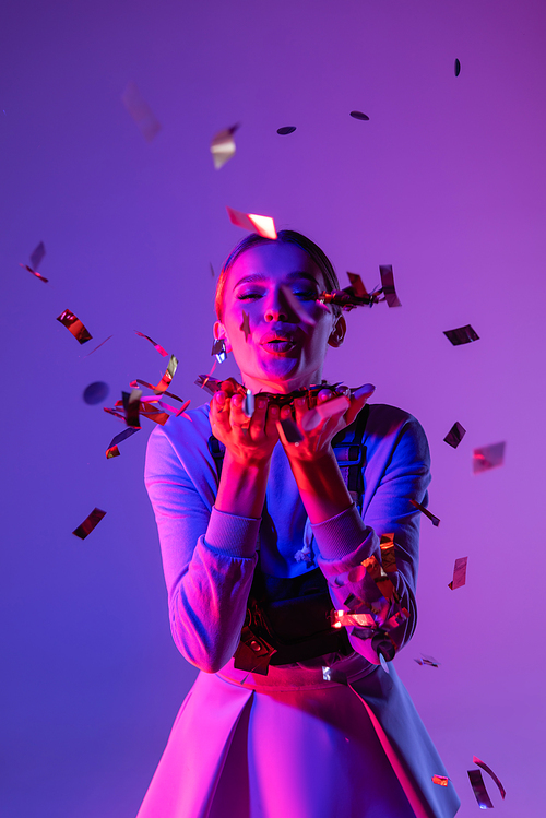 happy woman in stylish outfit sending air kiss near falling confetti on purple