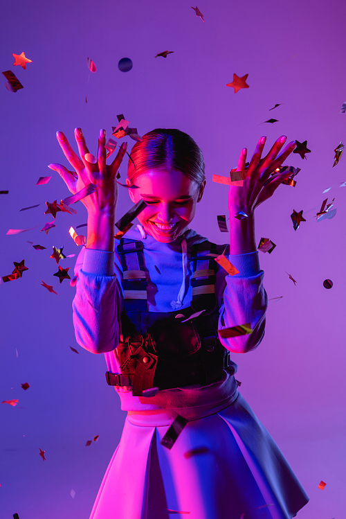 happy woman in stylish outfit throwing confetti on purple